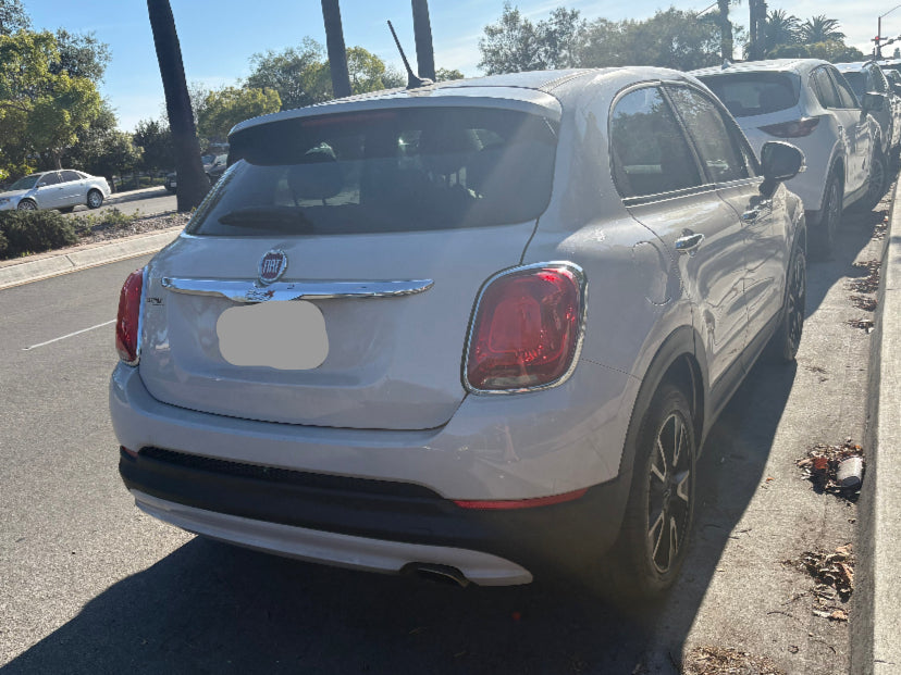 White car parked on the side of a street with trees and other cars in the background.